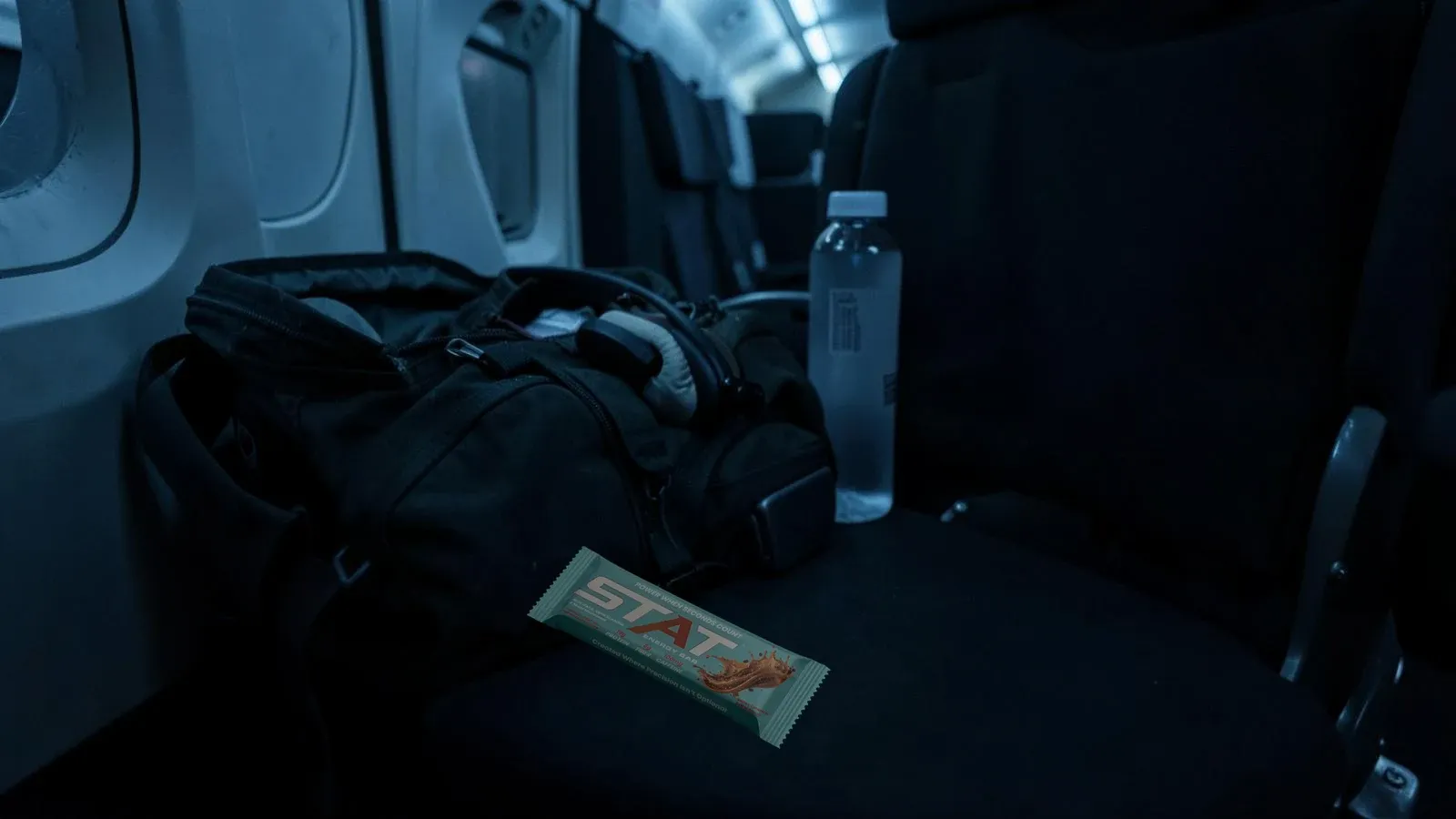 Dark documentary shot of a flight bag and headset on a crew rest seat with a real STAT Salted Caramel Coffee Energy Bar beside a water bottle under cool cabin light.